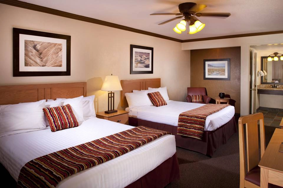 hotel room with 2 large beds, painting on the walls, a ceiling fan, and a small brown desk